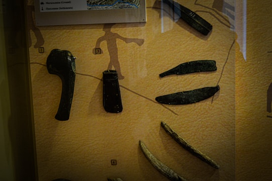 Various ancient tools and artifacts are displayed against a textured backdrop, with each item assigned a number for identification. The tools appear to be made of metal and stone, featuring axe heads, blades, and other implements. A silhouetted illustration of a human figure is visible in the background, adding context to the historical setting.