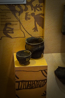 Two dark, textured ceramic vessels are displayed on a pedestal with a decorative pattern on its surface. The background features an abstract design with natural tones, resembling ancient artwork or engravings.