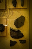 Various pieces of ancient pottery are displayed on a beige background. Each fragment features distinct textures and patterns, indicating they are part of historical artifacts. The pieces are mounted on plaques and arranged methodically, suggesting a museum exhibit.