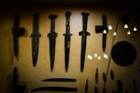 A display of ancient weapons, including several bronze daggers and other artifacts, set against a muted background. The objects have a patina indicating their age, and there are reflections of lights on the glass case.