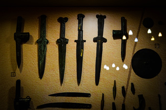 A display of ancient weapons, including several bronze daggers and other artifacts, set against a muted background. The objects have a patina indicating their age, and there are reflections of lights on the glass case.