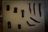 A collection of ancient, dark-colored tools or artifacts is displayed against a textured, beige background. The objects have various shapes, possibly made of metal, and are mounted on the surface for exhibition.