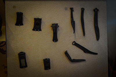 A collection of ancient, dark-colored tools or artifacts is displayed against a textured, beige background. The objects have various shapes, possibly made of metal, and are mounted on the surface for exhibition.