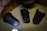 Three ancient, dark metallic tools or artifacts are displayed on a beige surface. They have flat, rectangular shapes with slightly raised edges, suggesting they might have been used for pressing or shaping. Each artifact has unique wear and markings, indicating age and use. The background features a pattern of black abstract symbols on a golden-yellow surface, and a large, weathered earthen jar is partially visible.