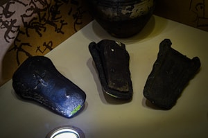 Three ancient, dark metallic tools or artifacts are displayed on a beige surface. They have flat, rectangular shapes with slightly raised edges, suggesting they might have been used for pressing or shaping. Each artifact has unique wear and markings, indicating age and use. The background features a pattern of black abstract symbols on a golden-yellow surface, and a large, weathered earthen jar is partially visible.