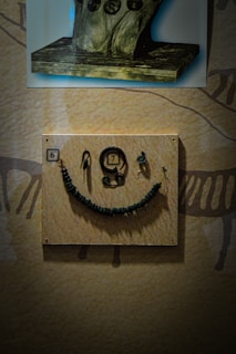 A display featuring ancient artifacts, including a panel with various items such as a beaded necklace and spiral-shaped objects. Above this, there is an image of a stone pedestal with engraved circular motifs.