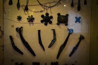A collection of ancient metal artifacts displayed on a textured surface, including various tools and abstract shapes. The items are dark and appear to be aged, arranged in rows for viewing.