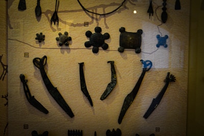 A collection of ancient metal artifacts displayed on a textured surface, including various tools and abstract shapes. The items are dark and appear to be aged, arranged in rows for viewing.