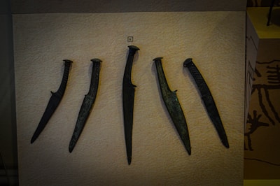 Five ancient daggers are displayed against a beige background. Each dagger has a distinct shape and design, with varying blade lengths and patterns on the handles.