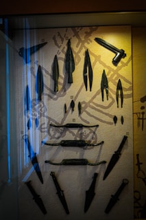 A collection of ancient and varied bladed weapons displayed vertically on a textured beige background, including daggers, swords, and spearheads. The display is enclosed in a glass case, with some reflections visible on the surface.