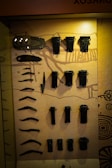 An assortment of black metallic artifacts is displayed against a warm beige background, possibly within a museum exhibit. The items vary in shape and size, including long, curved pieces and cylindrical objects, and appear to be organized neatly in rows. The background includes illustrative patterns and a light source casts a glow on the upper portion of the display.
