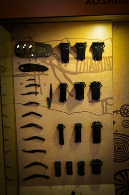 An assortment of black metallic artifacts is displayed against a warm beige background, possibly within a museum exhibit. The items vary in shape and size, including long, curved pieces and cylindrical objects, and appear to be organized neatly in rows. The background includes illustrative patterns and a light source casts a glow on the upper portion of the display.