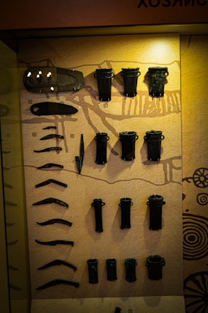 An assortment of black metallic artifacts is displayed against a warm beige background, possibly within a museum exhibit. The items vary in shape and size, including long, curved pieces and cylindrical objects, and appear to be organized neatly in rows. The background includes illustrative patterns and a light source casts a glow on the upper portion of the display.