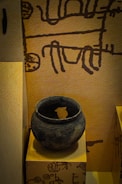 The image displays an ancient clay pot placed on a pedestal. The pot is round, dark, and has intricate carvings running around its surface. The background features a wall with primitive, abstract drawings in a dark color against a light brown surface.