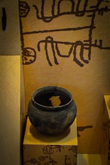 The image displays an ancient clay pot placed on a pedestal. The pot is round, dark, and has intricate carvings running around its surface. The background features a wall with primitive, abstract drawings in a dark color against a light brown surface.