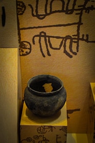 The image displays an ancient clay pot placed on a pedestal. The pot is round, dark, and has intricate carvings running around its surface. The background features a wall with primitive, abstract drawings in a dark color against a light brown surface.