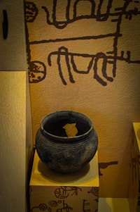 The image displays an ancient clay pot placed on a pedestal. The pot is round, dark, and has intricate carvings running around its surface. The background features a wall with primitive, abstract drawings in a dark color against a light brown surface.
