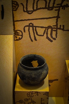 The image displays an ancient clay pot placed on a pedestal. The pot is round, dark, and has intricate carvings running around its surface. The background features a wall with primitive, abstract drawings in a dark color against a light brown surface.