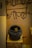The image displays an ancient clay pot placed on a pedestal. The pot is round, dark, and has intricate carvings running around its surface. The background features a wall with primitive, abstract drawings in a dark color against a light brown surface.