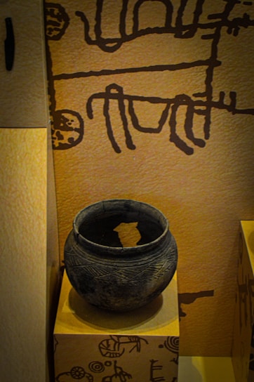 The image displays an ancient clay pot placed on a pedestal. The pot is round, dark, and has intricate carvings running around its surface. The background features a wall with primitive, abstract drawings in a dark color against a light brown surface.