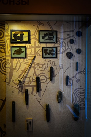 A museum exhibit showcasing ancient tools or artifacts mounted on a display board. Some items are enclosed in glass frames, while others are mounted directly onto the board. The backdrop includes illustrations or patterns that may resemble ancient maps or symbolic designs.