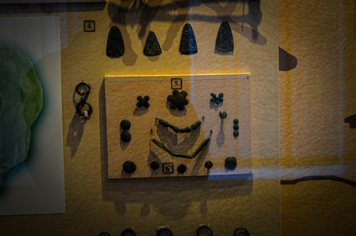 The image displays a museum exhibit showcasing various stone tools or artifacts, organized in a display case. There are several dark, irregularly shaped objects arranged in a symmetrical pattern, with numbers indicating the order or type of item displayed.