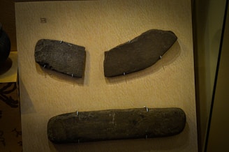 The image features three stone tools displayed on a beige board. Each tool is held in place by metal mounts. The tools vary in shape and size, with the top left and top right stones having more irregular shapes, while the bottom one is elongated and rectangular.
