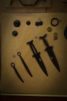 A collection of ancient artifacts displayed on a textured beige background. The objects include several bronze or iron implements such as daggers or small swords with dark patinas. There are also circular and disc-shaped items, possibly old coins or button-like objects. Each item is labeled with a small numbered tag for identification.