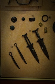 A collection of ancient artifacts displayed on a textured beige background. The objects include several bronze or iron implements such as daggers or small swords with dark patinas. There are also circular and disc-shaped items, possibly old coins or button-like objects. Each item is labeled with a small numbered tag for identification.