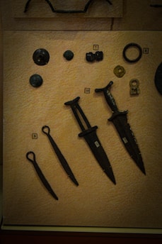 A collection of ancient artifacts displayed on a textured beige background. The objects include several bronze or iron implements such as daggers or small swords with dark patinas. There are also circular and disc-shaped items, possibly old coins or button-like objects. Each item is labeled with a small numbered tag for identification.