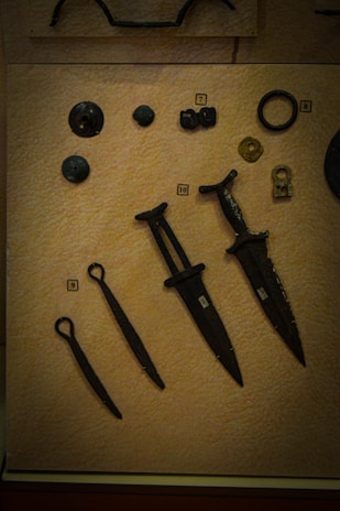 A collection of ancient artifacts displayed on a textured beige background. The objects include several bronze or iron implements such as daggers or small swords with dark patinas. There are also circular and disc-shaped items, possibly old coins or button-like objects. Each item is labeled with a small numbered tag for identification.