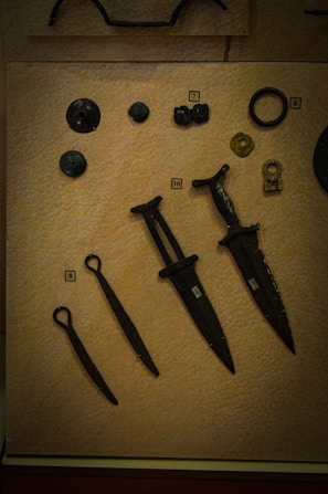 A collection of ancient artifacts displayed on a textured beige background. The objects include several bronze or iron implements such as daggers or small swords with dark patinas. There are also circular and disc-shaped items, possibly old coins or button-like objects. Each item is labeled with a small numbered tag for identification.