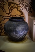 A dark, weathered clay pot with visible cracks and a rounded shape, placed on a light surface. In the background, there is a wall adorned with ancient-style petroglyph designs featuring figures and animals.