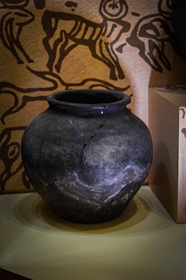 A dark, weathered clay pot with visible cracks and a rounded shape, placed on a light surface. In the background, there is a wall adorned with ancient-style petroglyph designs featuring figures and animals.