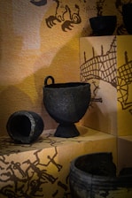 Several ancient pottery artifacts are displayed on a shelf, against a background with artistic illustrations. The pottery pieces appear to be made of dark, textured materials, possibly metal or clay, and vary in shapes and sizes including a bowl and a cup with a handle.