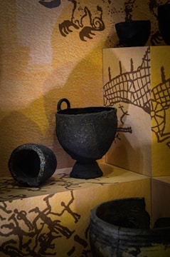 Several ancient pottery artifacts are displayed on a shelf, against a background with artistic illustrations. The pottery pieces appear to be made of dark, textured materials, possibly metal or clay, and vary in shapes and sizes including a bowl and a cup with a handle.