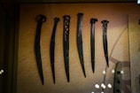 A display of six ancient dagger-like weapons with unique designs, each varying in length and handle style. The weapons are made of metal and are securely mounted on a textured background in a museum setting.