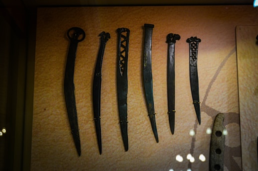 A display of six ancient dagger-like weapons with unique designs, each varying in length and handle style. The weapons are made of metal and are securely mounted on a textured background in a museum setting.
