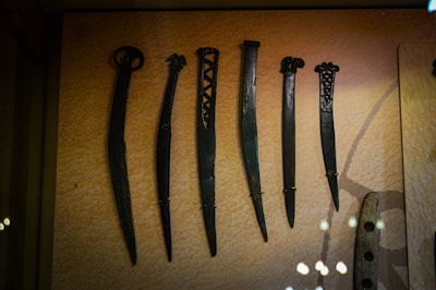 A display of six ancient dagger-like weapons with unique designs, each varying in length and handle style. The weapons are made of metal and are securely mounted on a textured background in a museum setting.