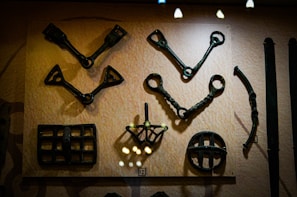 A display of various metal artifacts mounted on a wall, predominantly featuring tools or implements with intricate designs and geometric shapes. The items appear aged with a dark patina, suggesting historical significance.