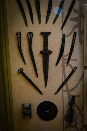An assortment of ancient metal artifacts displayed in a glass case, arranged symmetrically. These items include a central dagger or sword with a broad blade and a wide hilt, surrounded by smaller pointed tools or weapons. The artifacts appear to be made of a dark, aged metal, likely iron or bronze, and exhibit a rustic, weathered appearance indicative of historical significance.