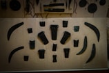 This is a display case showcasing various ancient tools or weapons arranged on a beige background. The items appear to be made of metal and have shapes resembling blades, axes, and chisels. The objects are symmetrically placed and vary in size and shape, with some being more curved and others being more rectangular.