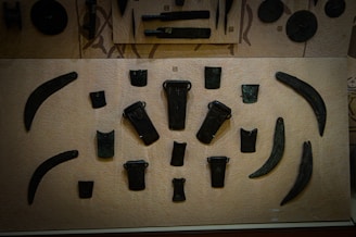This is a display case showcasing various ancient tools or weapons arranged on a beige background. The items appear to be made of metal and have shapes resembling blades, axes, and chisels. The objects are symmetrically placed and vary in size and shape, with some being more curved and others being more rectangular.