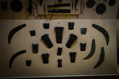 This is a display case showcasing various ancient tools or weapons arranged on a beige background. The items appear to be made of metal and have shapes resembling blades, axes, and chisels. The objects are symmetrically placed and vary in size and shape, with some being more curved and others being more rectangular.