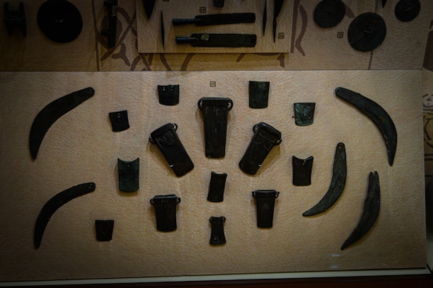 This is a display case showcasing various ancient tools or weapons arranged on a beige background. The items appear to be made of metal and have shapes resembling blades, axes, and chisels. The objects are symmetrically placed and vary in size and shape, with some being more curved and others being more rectangular.