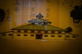 A detailed miniature model of an ancient Asian building with multiple layers and a tiled roof is displayed against a background featuring Chinese characters. The structure has a symmetrical design with small rectangular windows and an ornate central tower. The lighting creates a warm ambiance, highlighting the traditional architectural elements.