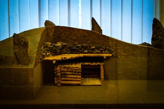 A miniature model of an ancient or traditional log cabin structure with a stone-covered roof set against a backdrop of vertical wooden panels. The model includes a roof made of stacked stones and walls constructed from small logs, giving a rustic and historical appearance.