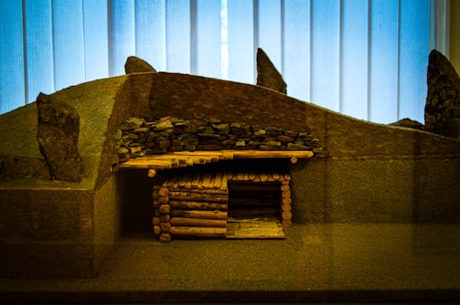 A miniature model of an ancient or traditional log cabin structure with a stone-covered roof set against a backdrop of vertical wooden panels. The model includes a roof made of stacked stones and walls constructed from small logs, giving a rustic and historical appearance.