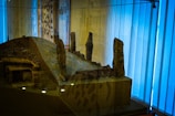 A scale model of an ancient structure is showcased in a glass display. It depicts a mound with several stone pillars standing upright. The model has a roofed section made of logs and is surrounded by a reflective glass case. Blue light filters through vertical blinds in the background, giving the scene a slightly serene ambiance.