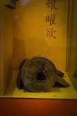 An ancient, round, metal object with inscriptions rests on a display stand. The background consists of a warm, yellowish wall with vertical Asian script.
